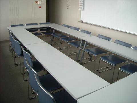 多数の机と青い椅子がコの字型に並べられ、部屋の奥にはホワイトボードが設置されている集会室Bの写真