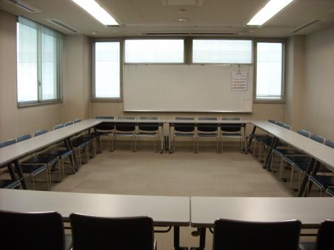 多くの長机と椅子がコの字型に配置されており、中央には空いたスペースがあり、部屋の奥にはホワイトボードと窓が確認できる集会室Aの写真