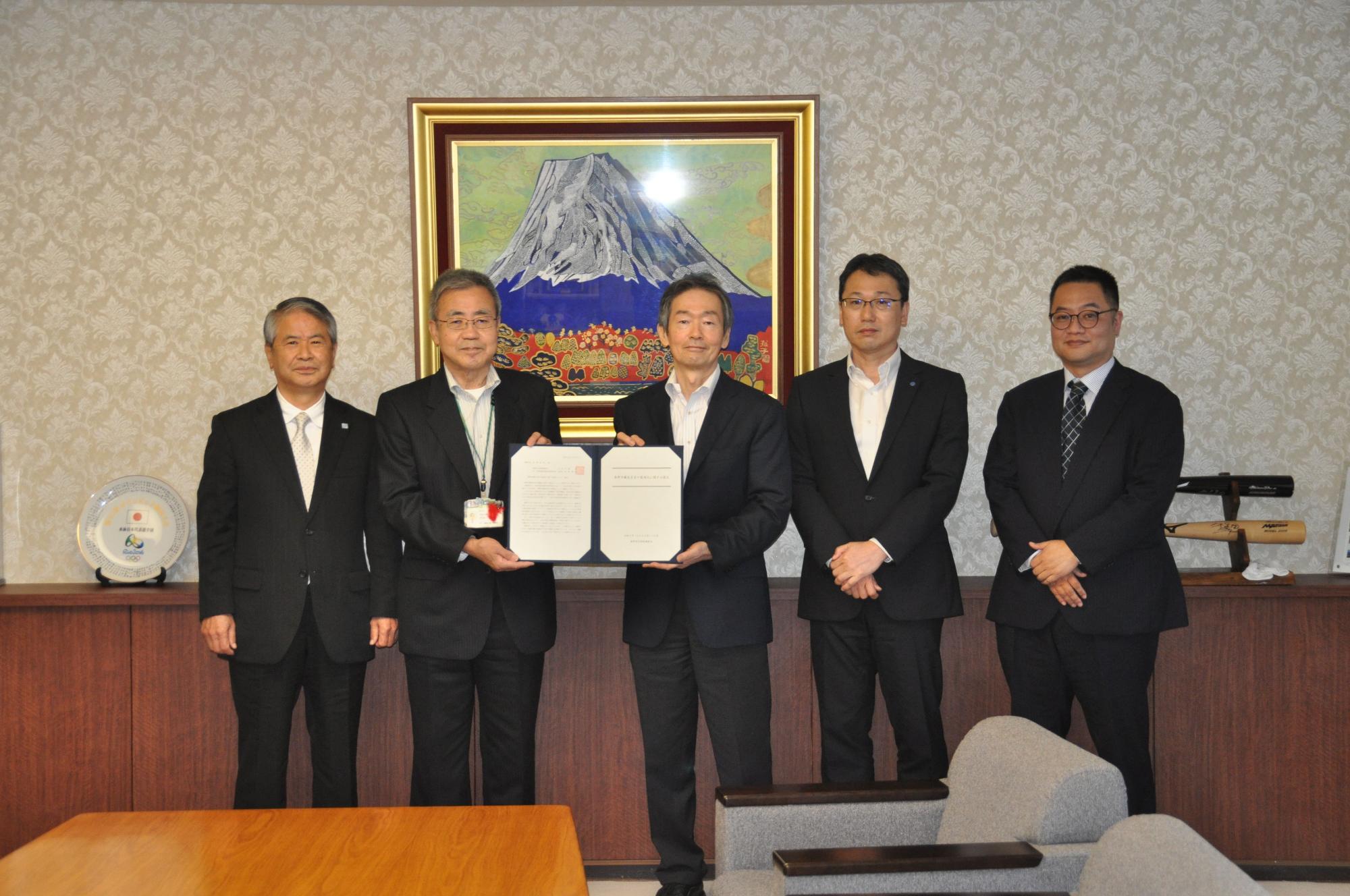富士山の絵画を背景に、黒いスーツ姿の5名の男性が並び、中央の作市長と板野部会長が報告書を掲げて記念撮影している調印式の写真