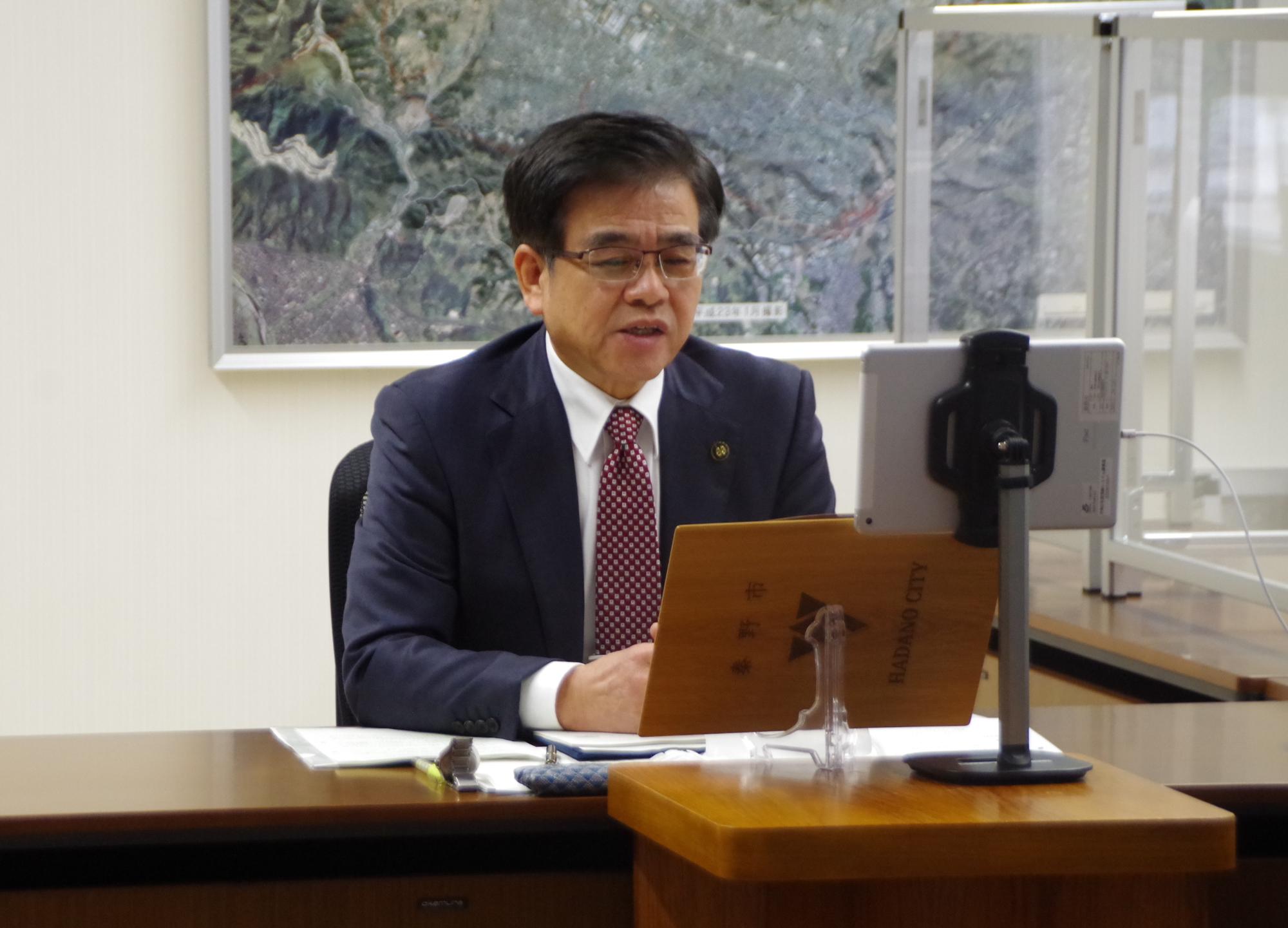 紺色のスーツに赤いネクタイを着用した市長が、デスクに設置されたモニターとノートパソコンを前にオンライン会議に参加している室内の写真