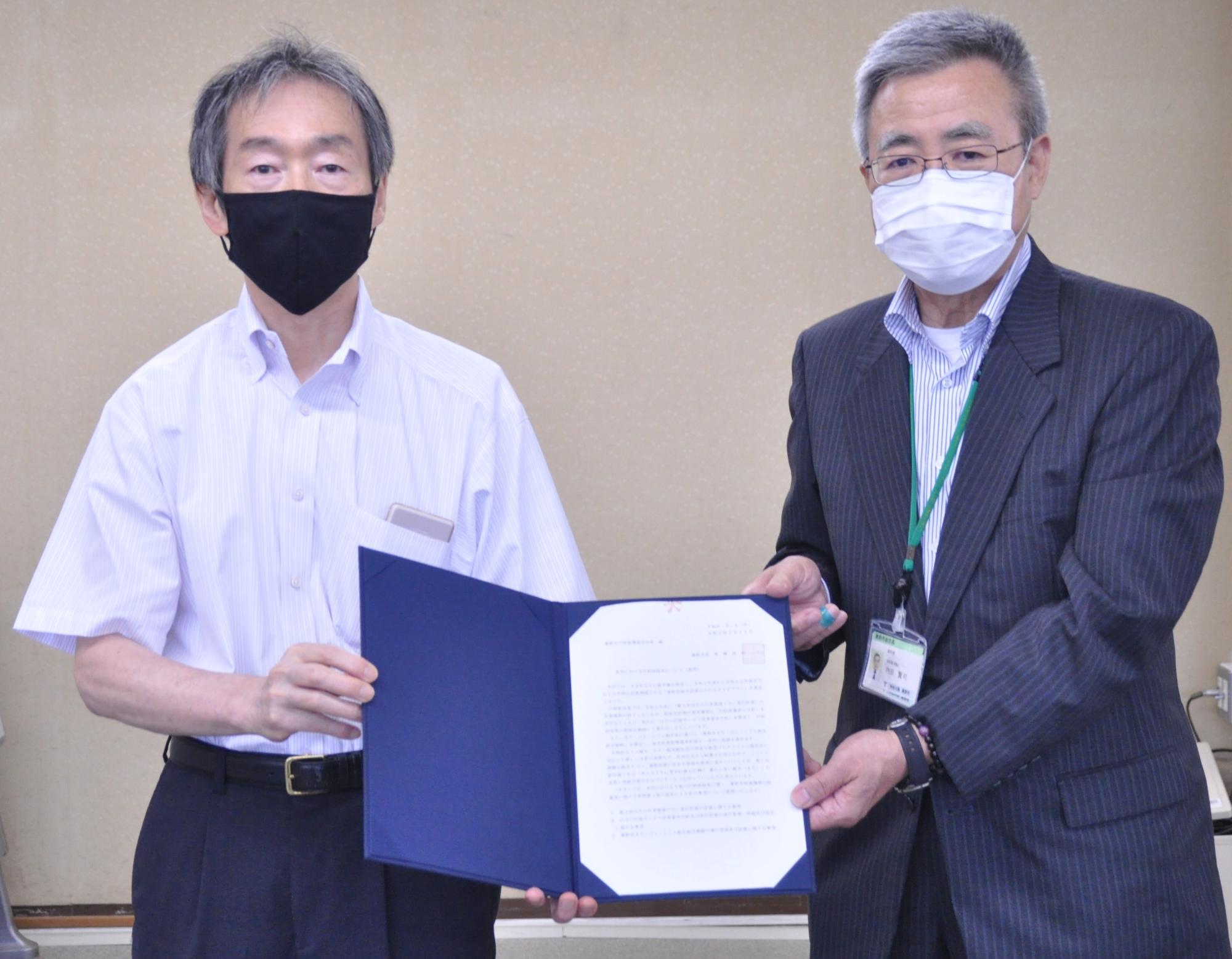 白と紺のピンストライプのスーツを着て白いマスクをつけた男性と、半袖の白いシャツに黒いマスクをつけた男性が、青いファイルに入った書類を並んで持ちながらカメラに向かって立っている写真