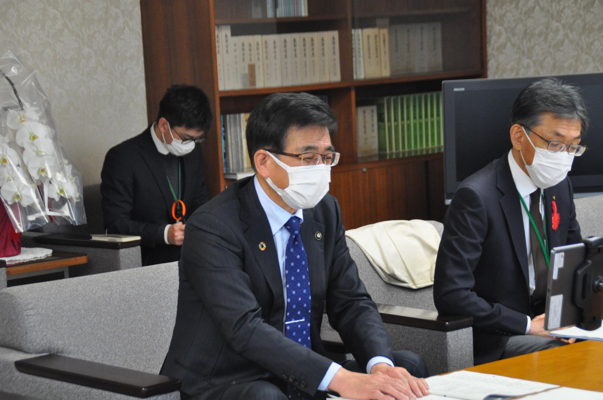 紺色のスーツに水玉模様のネクタイを着用した市長が、メガネと白いマスクをつけ、書類が広げられた木製のテーブルの前にあるソファに座っている様子の写真