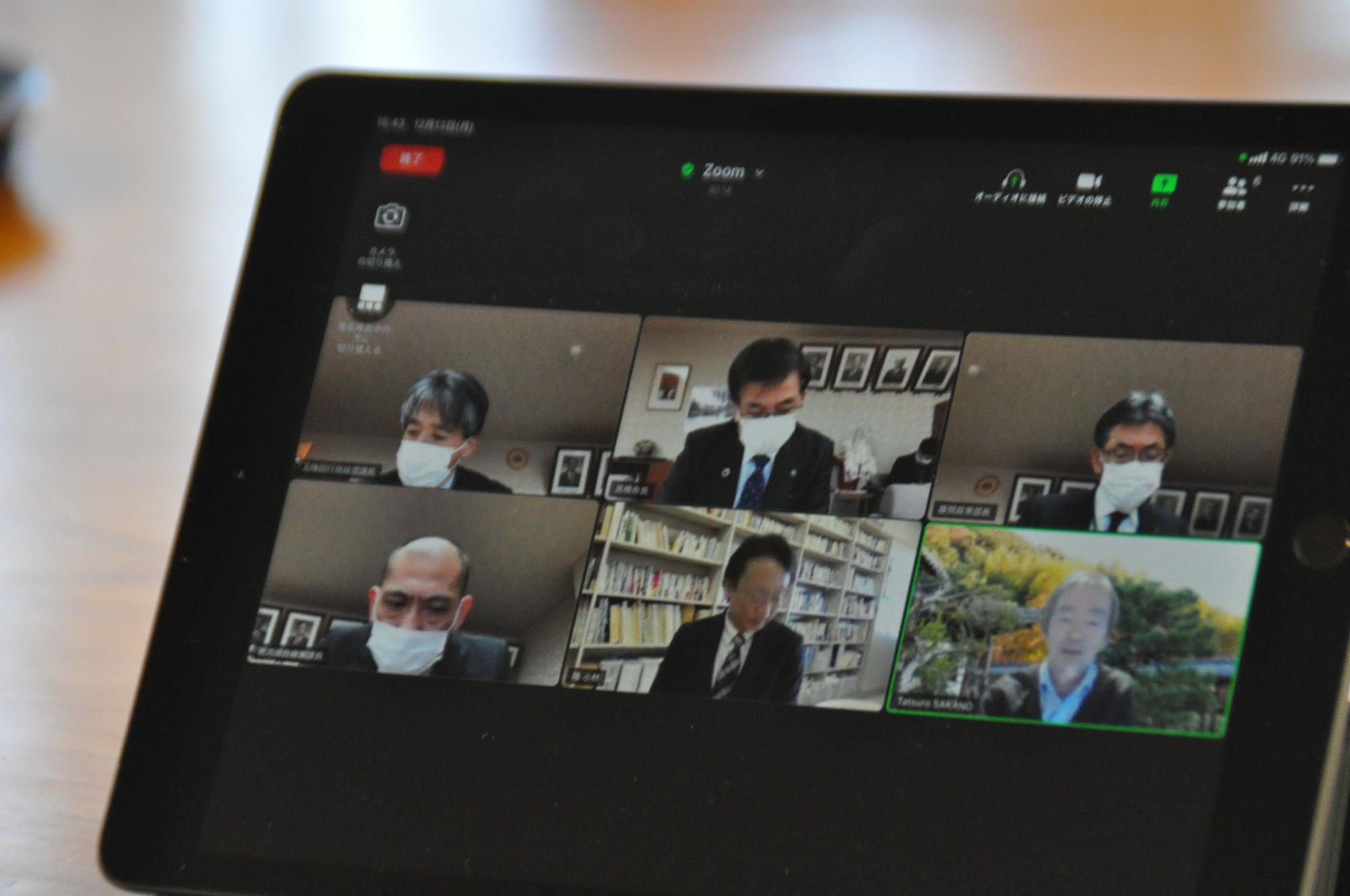 斜めから捉えられたタブレット端末の画面に、Zoomミーティングのグリッドビューが表示されており、白いマスクを着用した5人の男性と背景が屋外の男性1人が参加している様子の写真