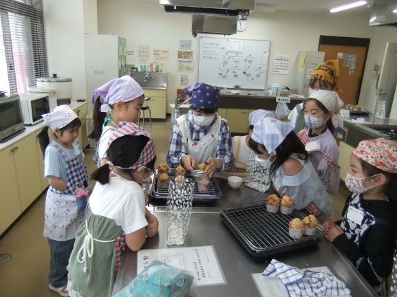 調理室で講師を周りに囲み子どもたちがカップケーキの作り方を教わっている