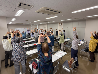 机を並べた部屋で大勢の大人が立って手を組んで背伸びしている
