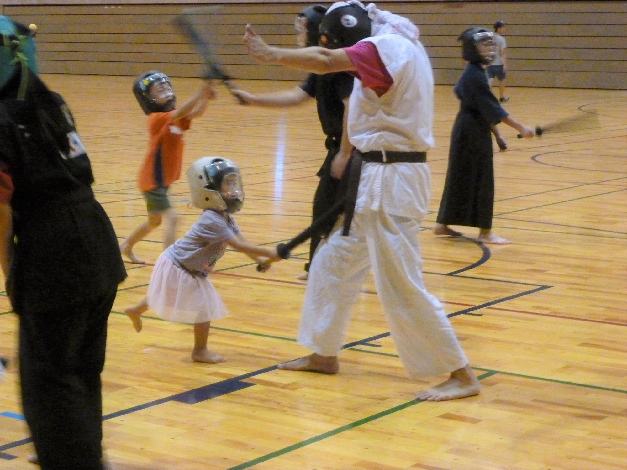 体育館のフローリングの床の上で、ヘルメットを着用した複数の大人が子どもたちに武術のような動きを教えており、特に白い道着姿の大人がピンクのスカートをはいた幼い女の子と竹刀のような棒を持って向かい合っている練習風景を捉えた写真