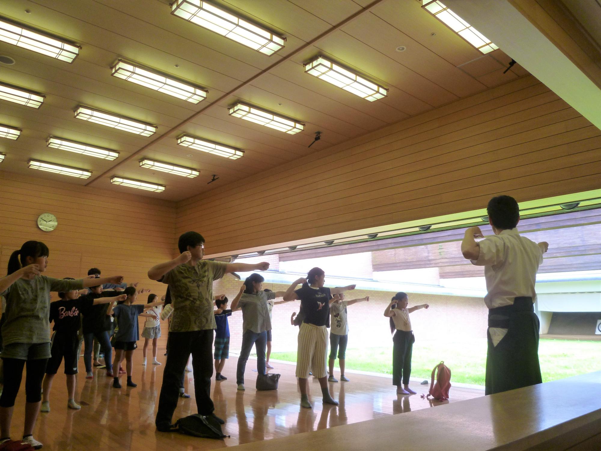 体育館のような木製の床と天井の明るい室内で、多くの参加者が一人の指導者に向かって腕を左右に広げる体操のような動きを一斉に行っている様子を低い位置から捉えた写真