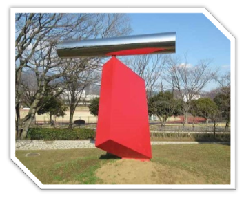 芝生広場の小高い丘に設置された抽象的なオブジェで、今にも倒れそうな斜めに設置された赤い多角形の土台の上に、銀色に輝く円筒形の物体が乗っている屋外彫刻の写真