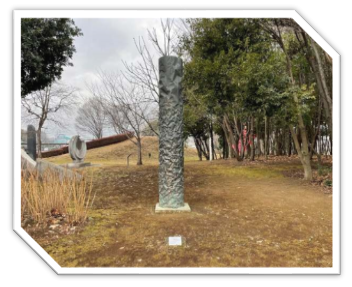 屋外の木々に囲まれた芝生の上に立つ、黒っぽい円柱形の彫刻で、表面はごつごつとした岩肌のような質感を持つアート作品の写真