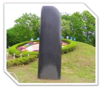 木々に囲まれた小高い広場の花壇や生垣で縁取られた芝生の上に立つ、黒く滑らかな表面を持つ、巨大な短刀のような形をした抽象的な彫刻の写真