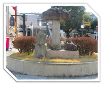 歩道の中央にある円形の植え込みにある長方形の土台に、顎に両手を置き物思いにふけっているような人物の石像と、雲のような形をした柱の彫刻、そして動物の彫刻が設置されている写真