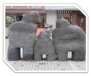 凹凸のある灰色がかった表面を持つ、親子を模したような三つの抽象的な石の彫刻が、商店街の歩道に寄り添うように設置されている写真