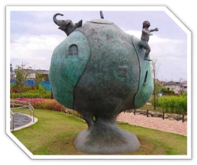 通路沿いの芝生広場に設置された、地面に土台とくびれのある大きな円形の地球のような形をし、表面に小さな窓がある抽象的な青銅色の彫刻の上に、ゾウや両手を前に伸ばしている躍動感のある子どもが配置された、物語性のある屋外彫刻の写真