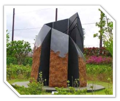 フェンスに囲まれた緑豊かな広場に設置された、長方形の表面がゴツゴツした様な茶色い石材が組み合わされた上から黒い液体を流したように、右側から左上に向かって螺旋状に配置された金属板が頂上に向かって尖るようにデザインされた、抽象的な屋外彫刻の写真