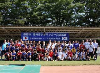屋外のグラウンドで、「秦野市・坡州少年サッカー交流試合」の横断幕が掲げられており、ユニフォーム姿の子供たちや関係者が、集合している写真