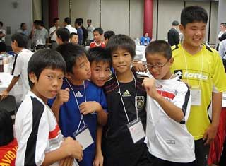 サッカーユニフォームまたはTシャツを着用した小学校高学年から中学生くらいの少年たちが、首から下げた名札を下げており、笑顔で肩を並べている写真