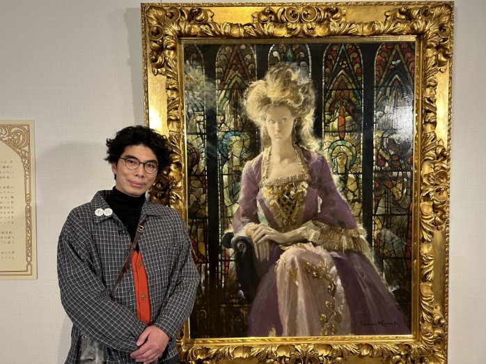 美術展の会場で、チェック柄のシャツを着た俳優の片桐仁氏が、豪華な額縁に入ったドレスを着た女性の肖像画の前に立っている写真。