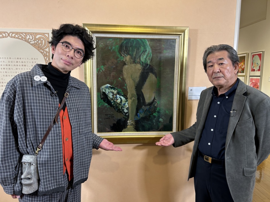 展示会の会場で、男性と俳優の片桐仁氏が背中が大きく開いた服を着た女性の肖像画の間に立ち作品を手で指し示して並んでいる記念写真