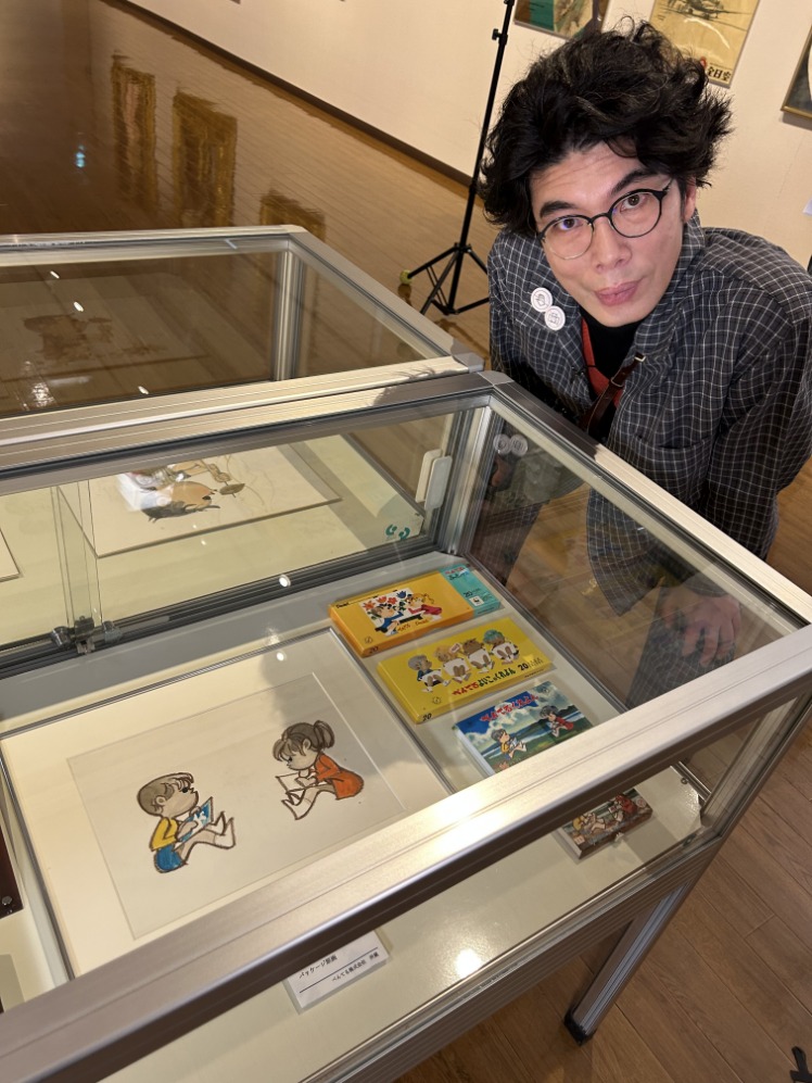チェック柄のシャツを着た俳優の片桐仁氏が、展示されたぺんてるの原画とクレヨンが並んでいるガラスケースの前に腰をかがめて立っている写真
