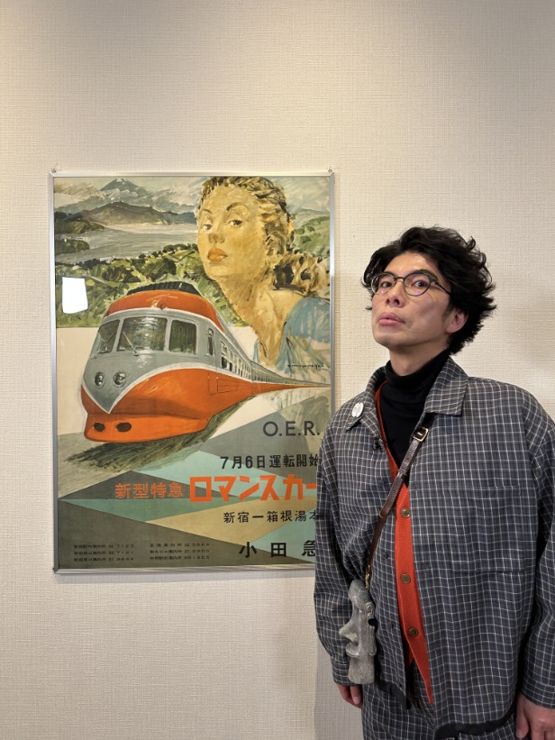 チェック柄のシャツを着た俳優の片桐仁氏が、オレンジと白のボディのロマンスカーや、女性の顔、風景などが描かれた「新型特急 ロマンスカー」と書かれたポスターの横に立っている写真