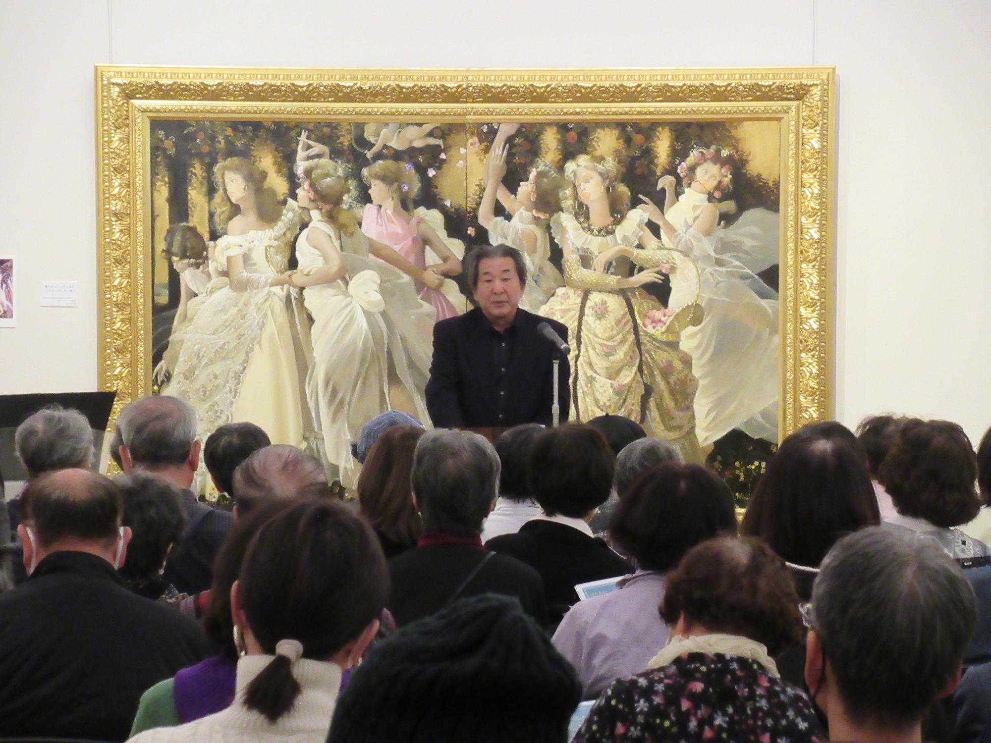 豪華な衣装を着て踊る大勢の女性や天使が描かれている絵画を背景に、マイクスタンドの前に立つ黒い服を着た男性が椅子に座った参加者に話しかけている写真