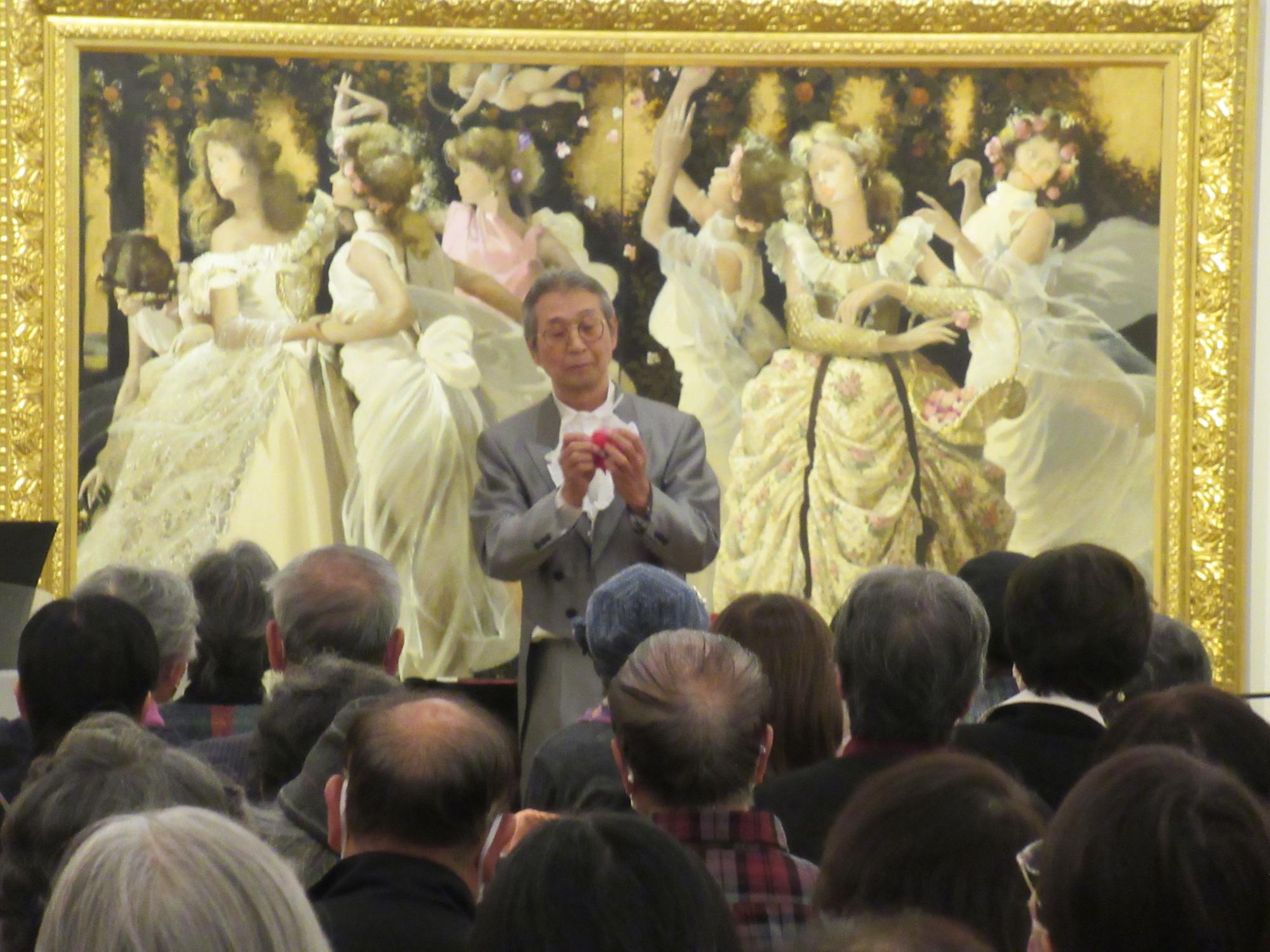 大勢の女性や天使が描かれた絵画を背景に、マイクスタンドの前に立ち、赤い花のようなものを両手で持って椅子に座っている参加者に話しているグレー色のジャケットを着た男性の写真
