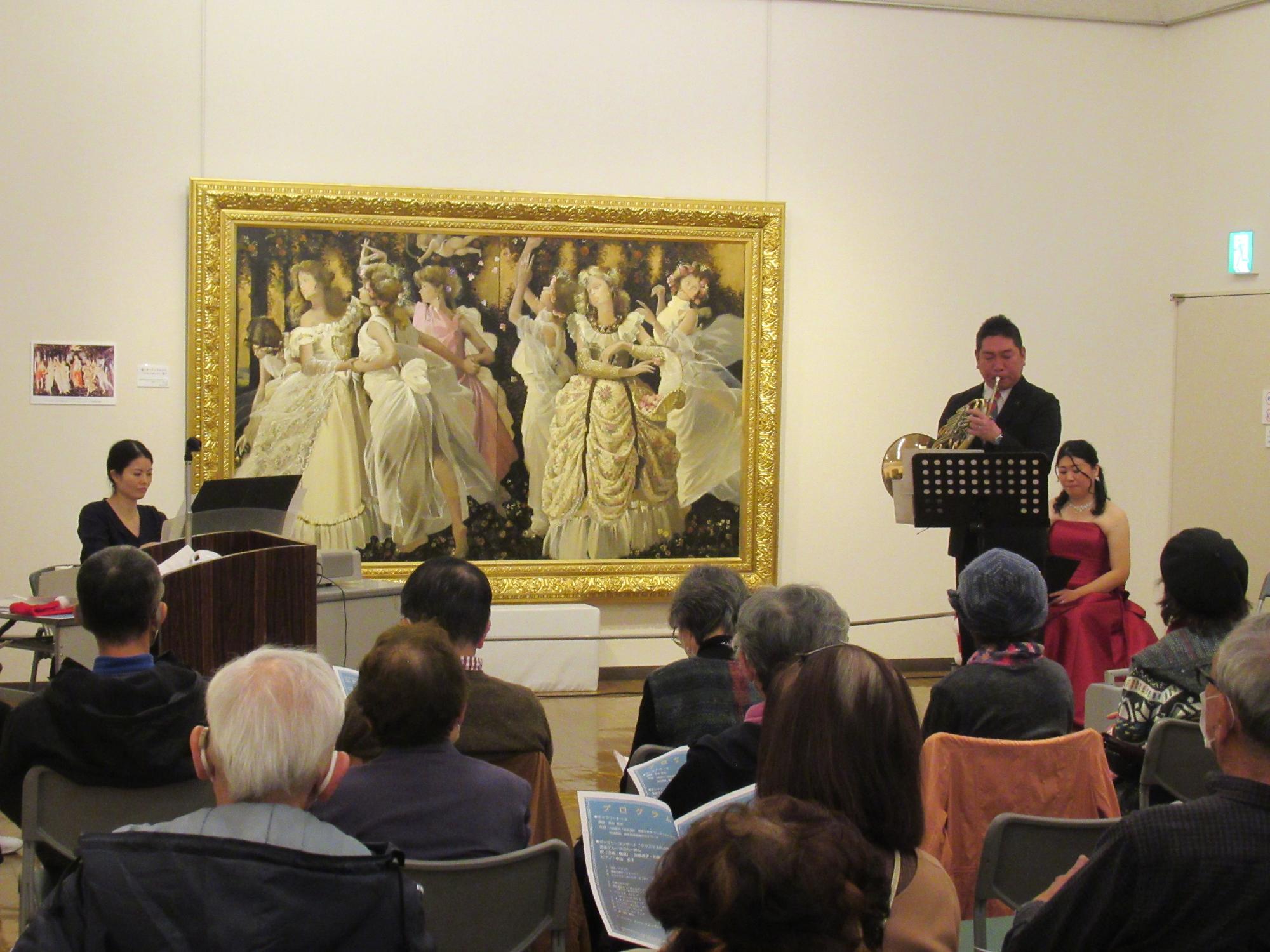 金色の額に入った絵画の前で、左側で黒色の服を着た女性がエレクトーンを演奏し、右側に立つ黒いスーツを着た男性がホルンを演奏し、後ろの椅子に座る赤いドレスを着た女性と、演奏会を鑑賞する人々が椅子に座って演奏を聞いている様子の写真