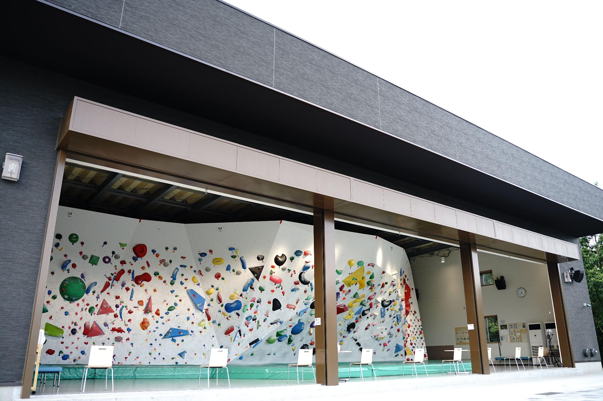 黒い外壁の建物の中に、白い椅子が等間隔で設置されており、カラフルなホールドが白い壁一面に取り付けられたクライミングジムの写真