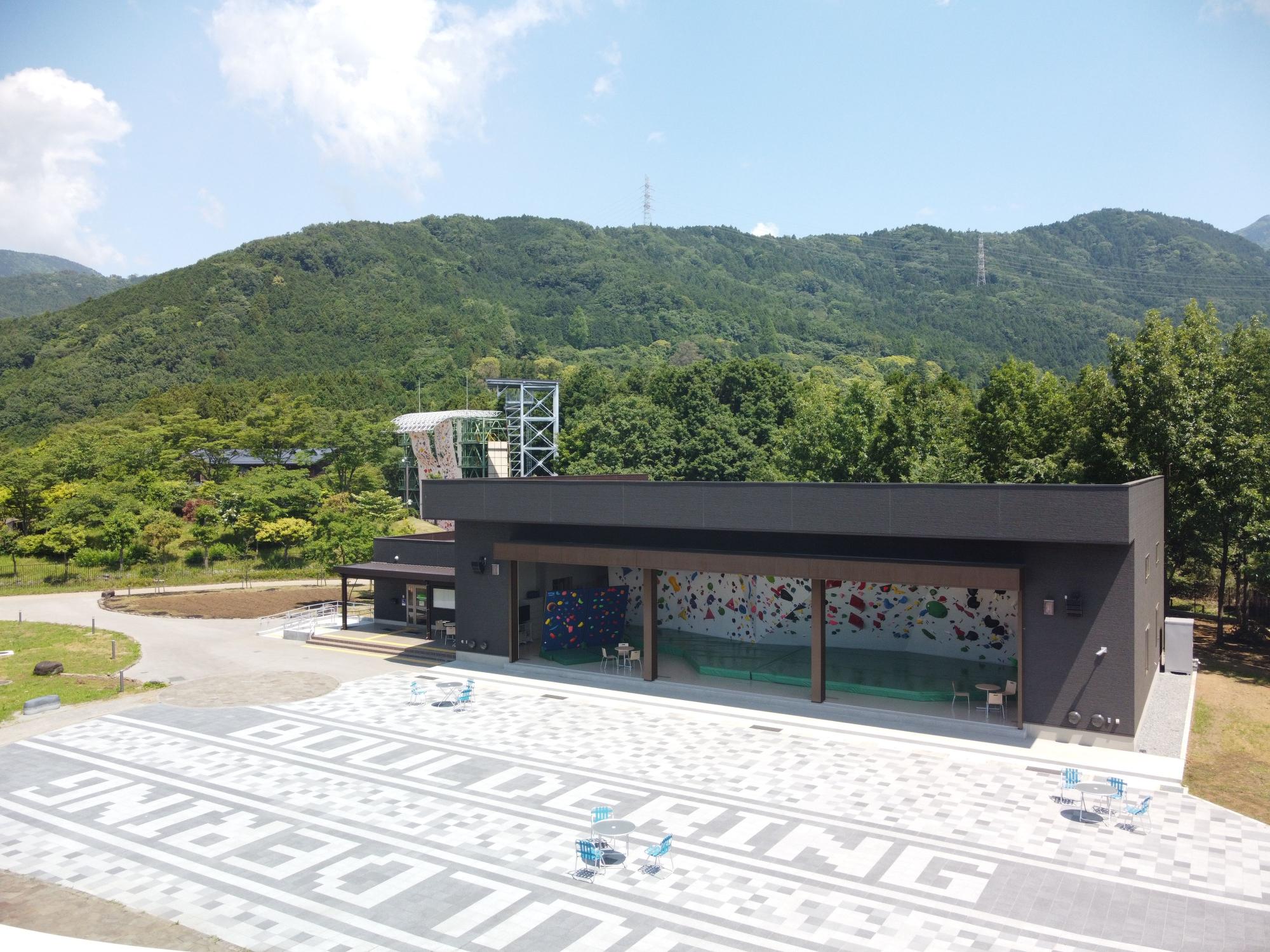 背景に山が見える景色で、建物の大きな開口部からはカラフルなクライミングウォールが見え、屋外の広場には3脚の椅子がセットされた3台のテーブルが設置されている写真