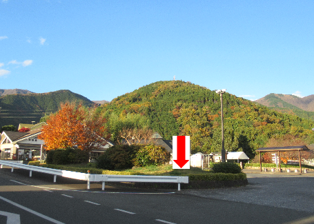 背景には緑豊かな山々が広がり、道路とロータリーの奥に、色とりどりの紅葉した木々や建物があり、敷地入り口のガードレール付近に赤い下向きの矢印が描かれている写真