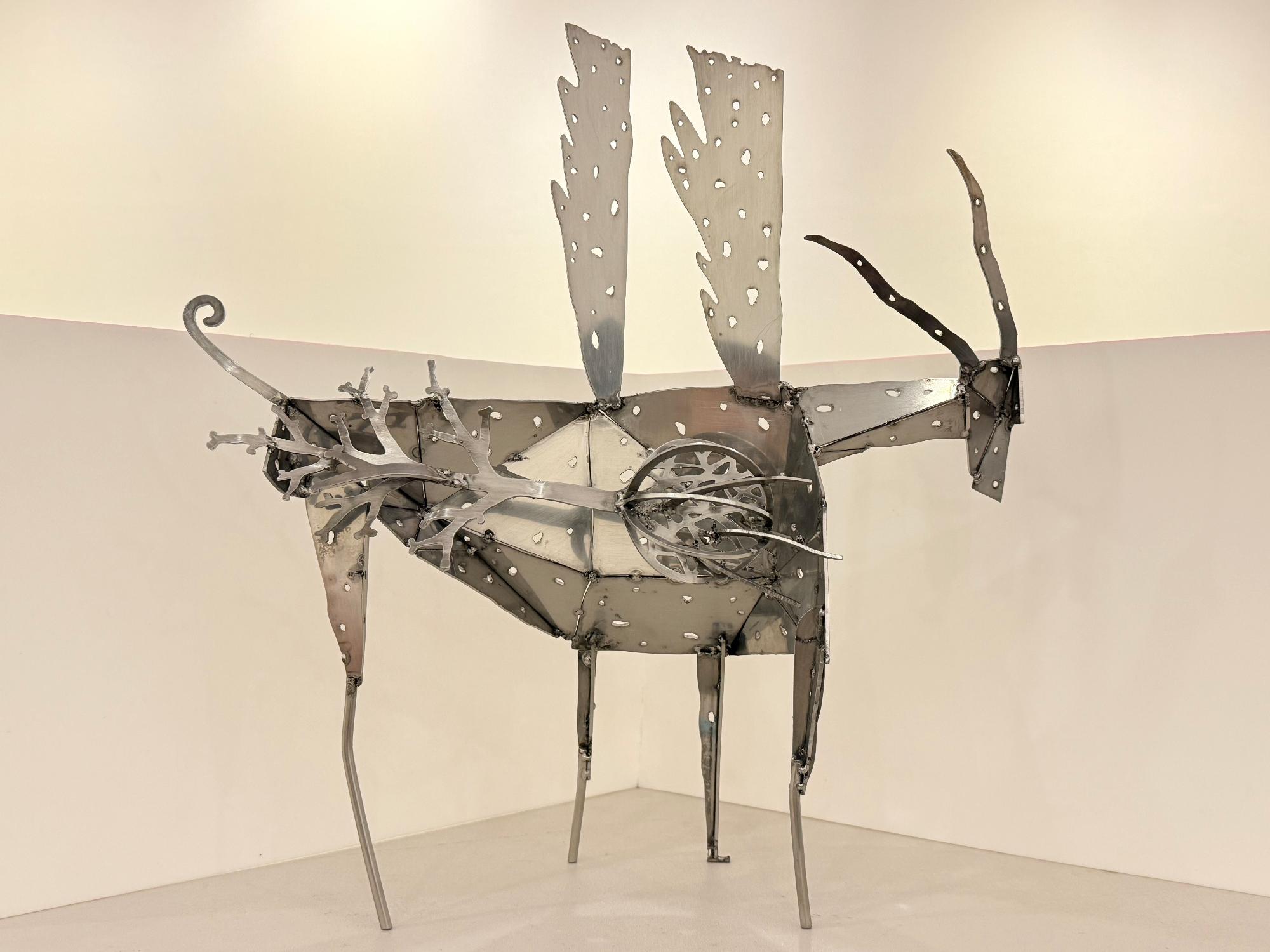 2つの翼や体全体に穴が開き、尻尾があり、ねじれた2本の角を持つ金属製の鹿を象ったモニュメントが、細長い四本の脚で立っている作品の写真