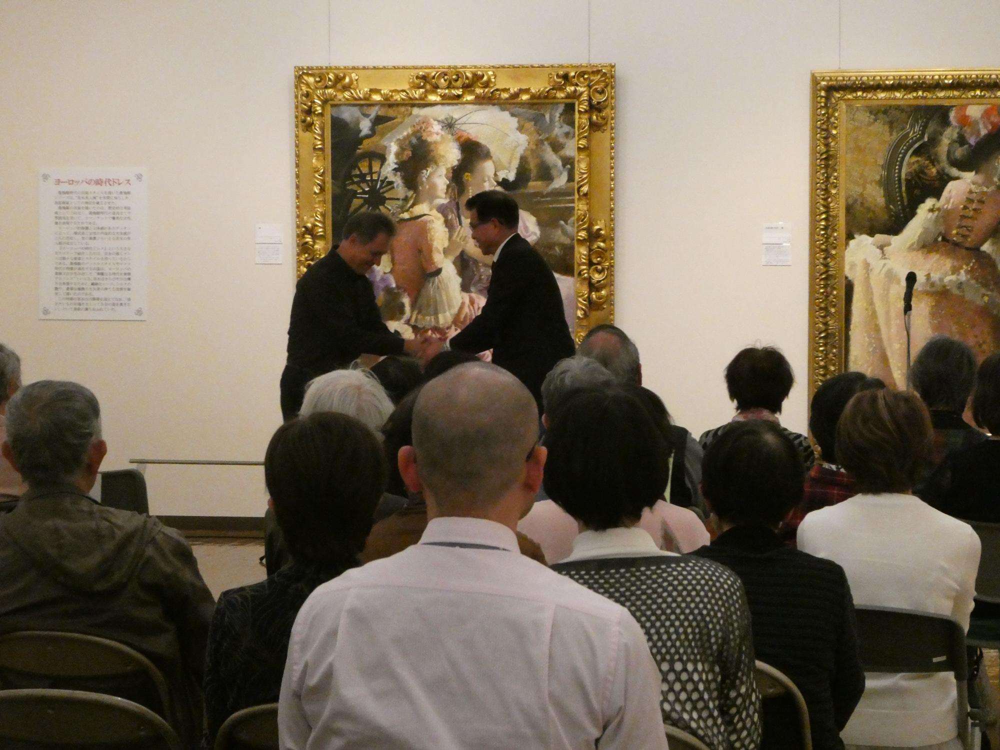 豪華な額縁に入った肖像画が展示された美術館で、多くの聴衆が椅子に座って前方を向く中、二人の男性が握手を交わしている様子の写真