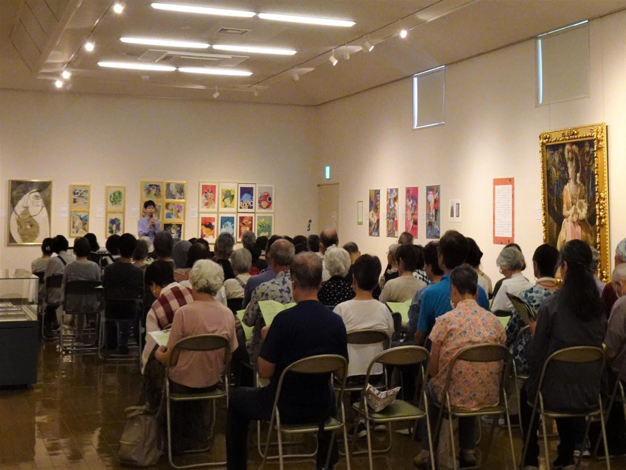 壁に沿って絵画やイラストが展示されている美術館で、多くの人々が椅子に座って前方を向き、ライトブルーのシャツを着た男性が聴衆に向かって話をしている様子を捉えた写真