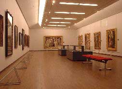 壁沿いに額縁に入った絵画が並び、中央には展示ケースと赤い長いベンチが配置された美術館の展示室全体の様子を写した写真
