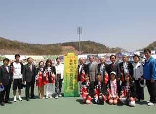 屋外のトラックで、秦野市民体育大会と書かれた看板を中心に、スーツや赤い法被、運動着を着用した男女が、整列し記念撮影している写真