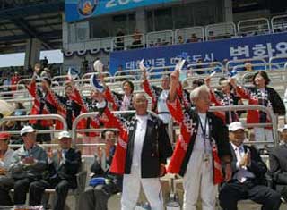 屋外の競技場の観客席で、複数の男女が赤と黒の法被を着用し、手に旗を持っており、後ろの方には韓国語で書かれた青い横断幕がある写真