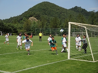 緑の芝生のサッカー場で、白いユニフォームと濃い色のユニフォームを着た少年たちがゴール前でサッカーボールを追いかけ、ゴールキーパーがシュートを止めようとしている試合中の写真