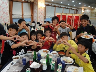 宴会場で、テーブルを囲みサッカー少年たちが、数人でハートマークを両手で作って笑顔を見せており、和やかな雰囲気で交流している様子の写真