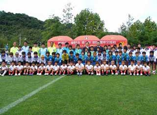 緑の芝生のサッカーグラウンドで、子供たちと指導者らしき大人たちが、ユニフォーム姿で整列し集合している写真