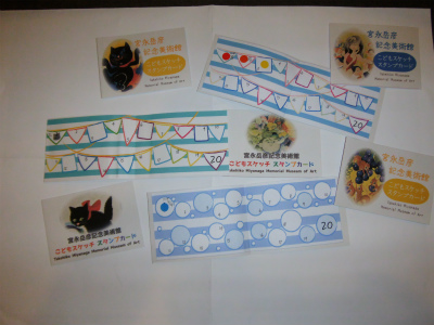 カラフルな旗や丸い模様が描かれたスタンプカードが3枚と、絵画や黒猫のイラストが描かれたカードが置かれている、美術館のスタンプカードの写真