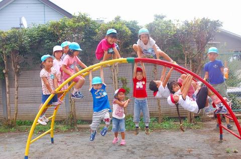 複数の子供たちが、赤と黄色に塗り分けられた半円形の太鼓はしごの遊具で、上段に座ったり、鉄棒のようにぶら下がって遊んでいる写真