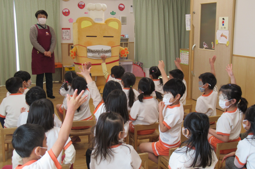 マスクを着用した園児たちがオレンジ色の椅子に座り、ボンチーヌが立っている前で、数人の園児が元気よく手を挙げている様子の写真