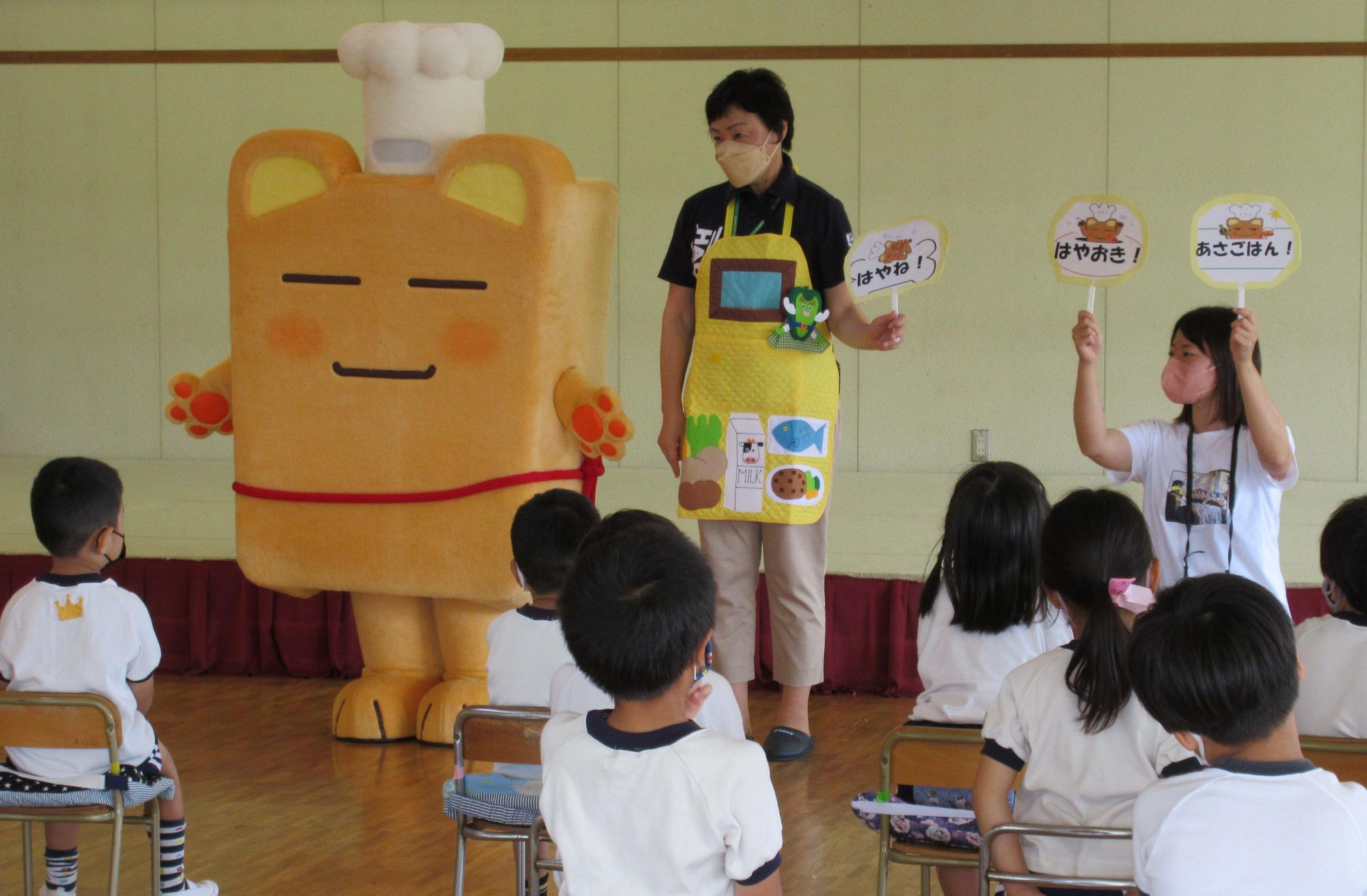 ボンチーヌと、エプロン姿の先生、そして「あさごはん!」などと書かれたプラカードを持つもう一人の先生が、床に座った園児たちに向けて、食育指導を行っている様子の写真