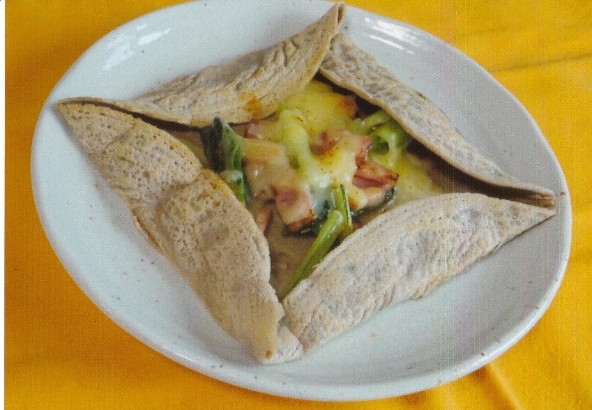 小松菜やベーコンの上にとろけたチーズがこんがり焼かれた生地の上にのっており、生地の外側が四つ折りに包まれた、正方形のガレットが白い皿に盛られている写真