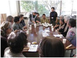 広い窓のある明るい部屋で、数人の高齢者が大きな円卓を囲んで座り、白衣を着た女性スタッフから料理を受け取ったり説明を聞いたりしている賑やかな集まりの様子の写真