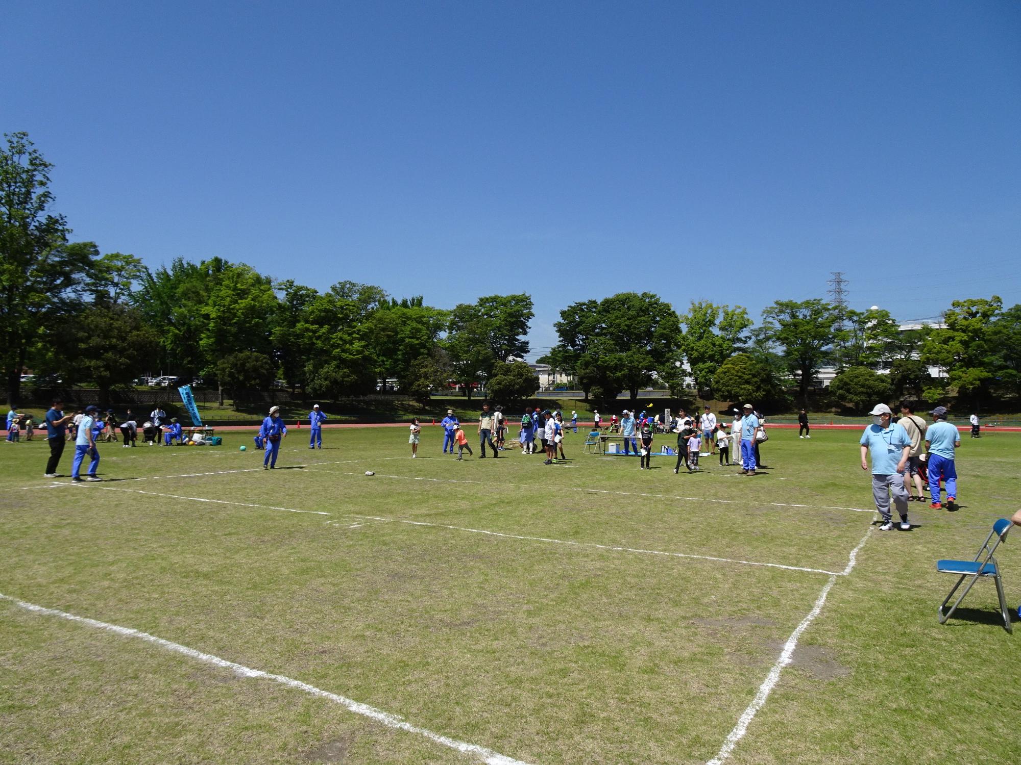 青空の下、広々とした芝生の広場で、子供から大人までスポーツを楽しむ人々が点在し、和やかな雰囲気の中で活動している様子の写真