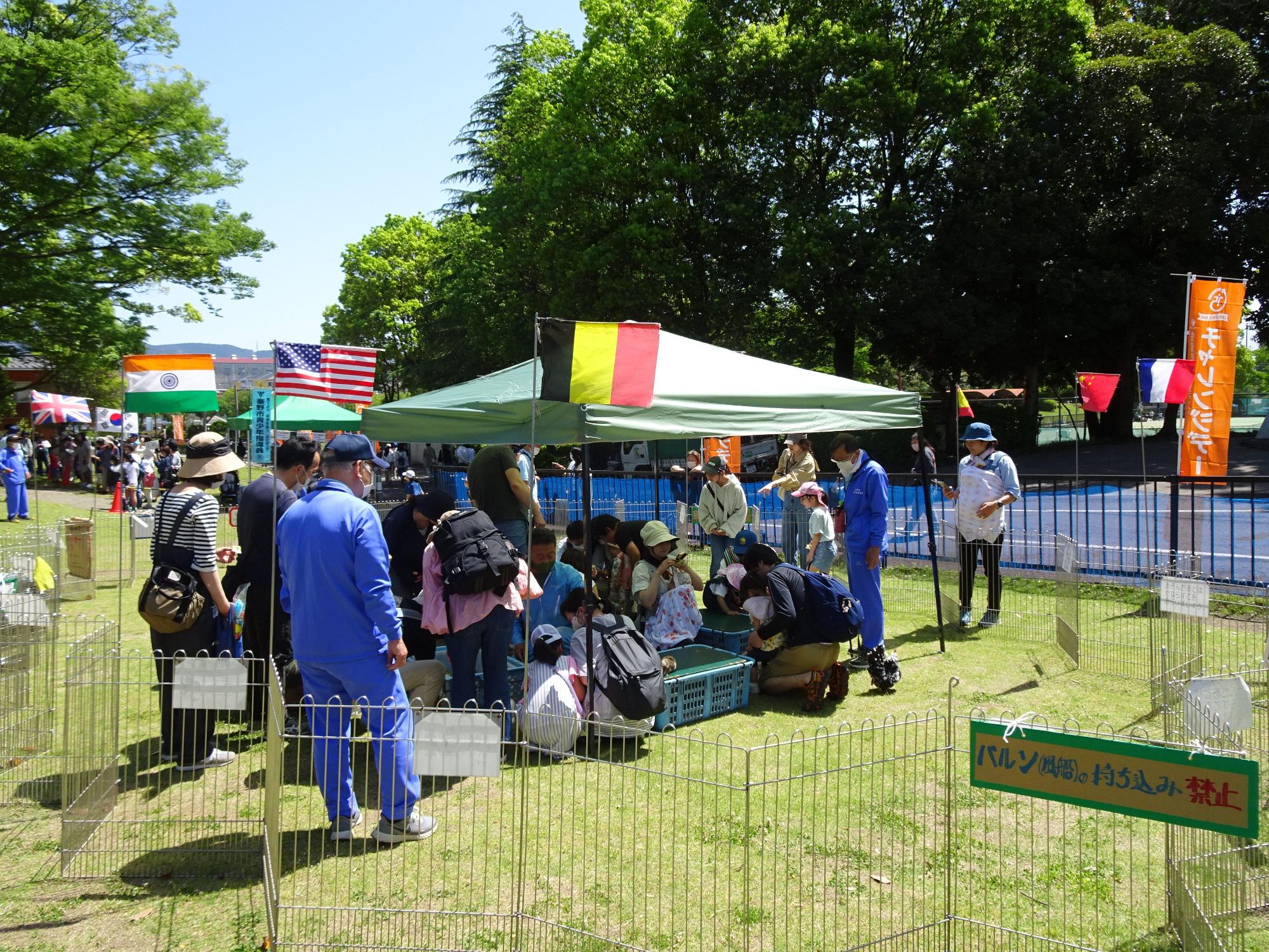 柵で囲まれた芝生の広場にテントが設置されており、そのテントの下に複数の子供や家族連れが小動物と触れ合っている写真