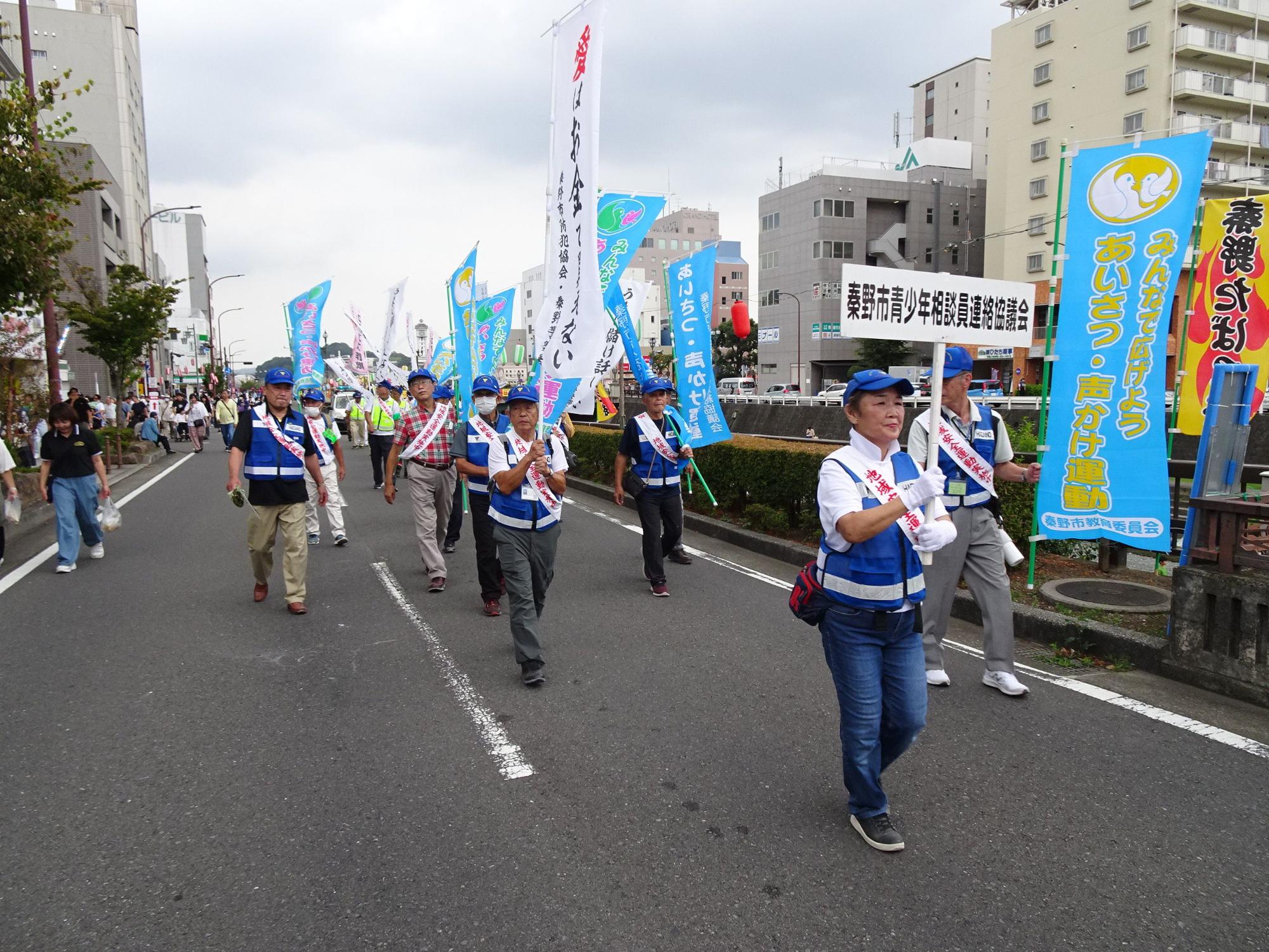 街中を歩行者天国にして行われている啓発パレードの様子で、青いキャップと青いベストを着用し、のぼり旗やプラカードを掲げた多数の市民活動家が、都市のビル群を背景に隊列を組んで道路を行進している写真