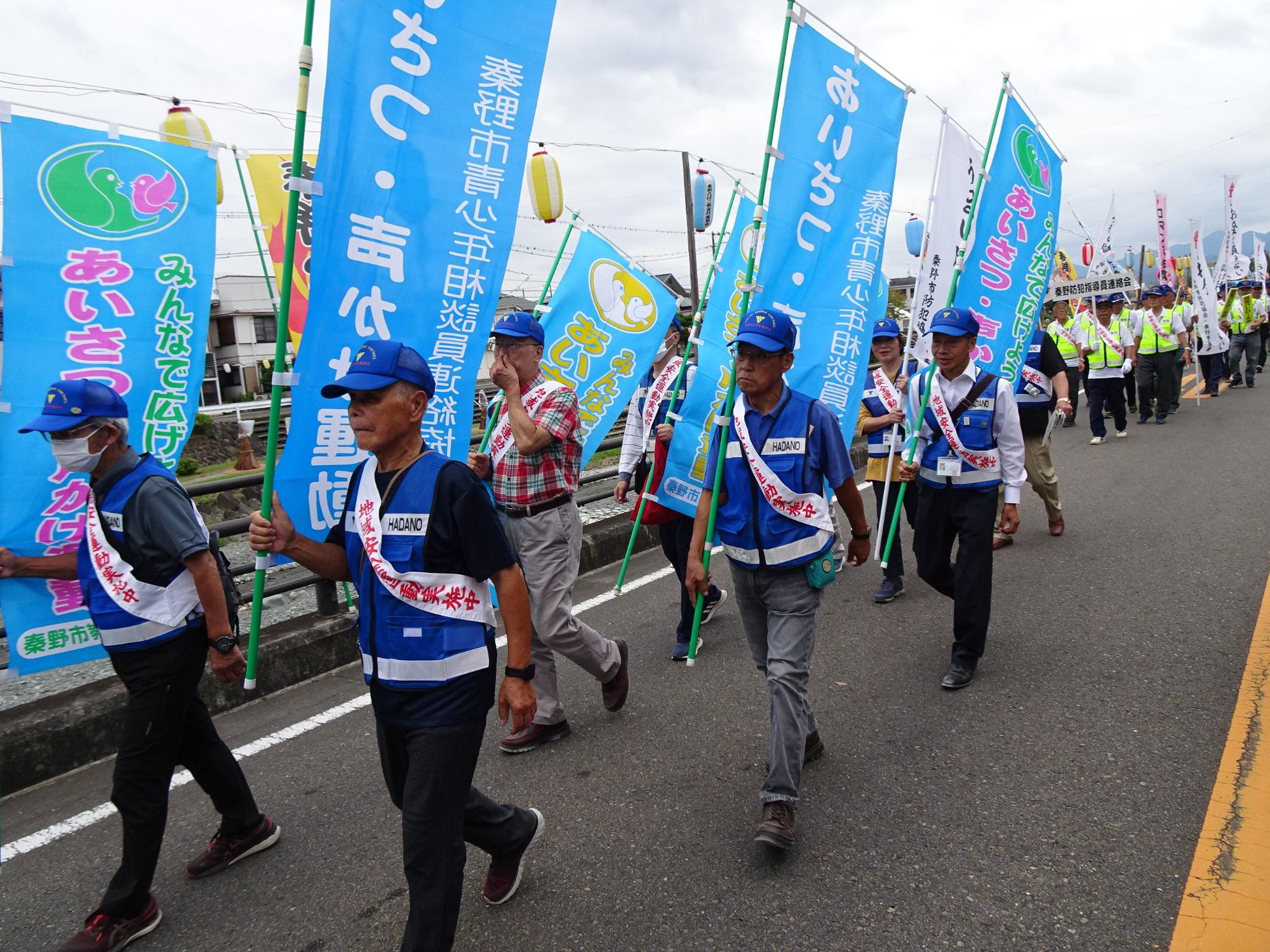 曇り空の下、青いベストとキャップを身に着け、秦野市のスローガンである「あいさつ・声かけ」と書かれた青いのぼり旗を掲げた市民活動家たちが、横断幕のようなものをくぐりながら集団で道路をパレードしている様子の写真