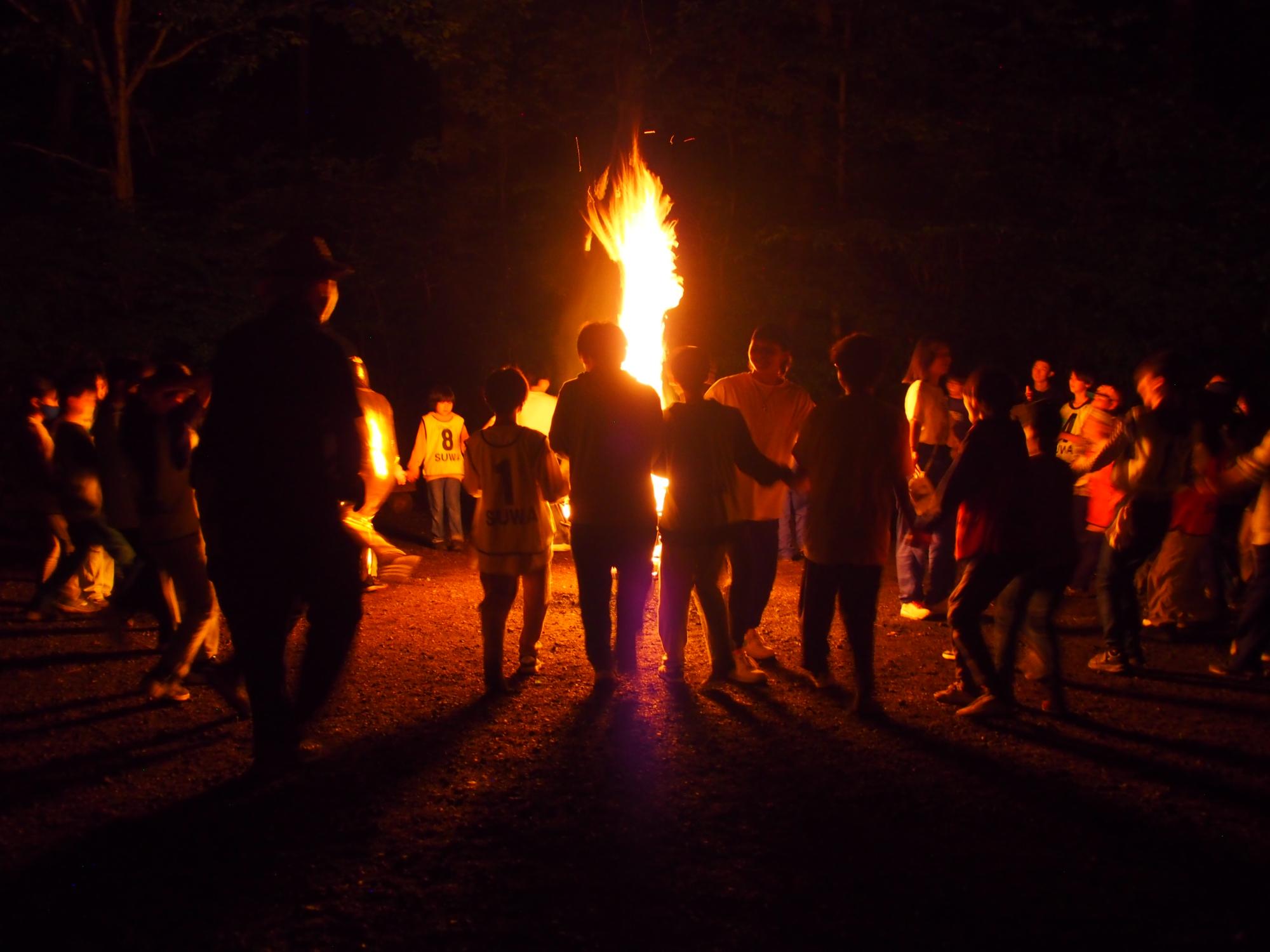 夜空の下、複数の子供や大人たちがキャンプファイヤーを囲み楽しんでいる写真